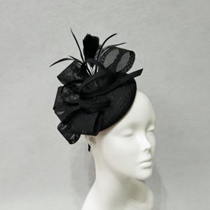May include: A black fascinator hat featuring a circular base, structured mesh loops, and upright black feathers. The headpiece is displayed on a white mannequin head, showcasing the intricate woven texture and layered design of the fabric elements.