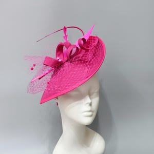 Könnte beinhalten: Ein leuchtend fuchsiafarbener Fascinator mit breiter, geschwungener Krempe, einer strukturierten Schleife und feinem Netz. Der Kopfschmuck ist mit dünnen, aufrechten fuchsiafarbenen Federn und kleinen, passenden kugelförmigen Akzenten verziert.