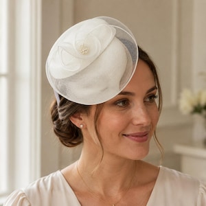 May include: A white fascinator hat featuring a circular base, a layered fabric flower accent at the centre, and a cluster of small white faux pearls. The headpiece is partially covered with a fine white netting veil that extends over the edges.