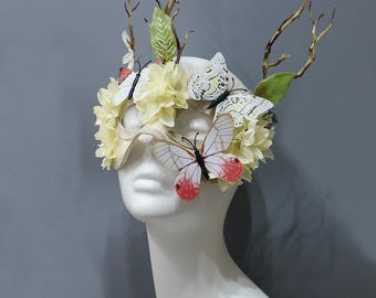 May include: A decorative mask with a butterfly and floral design. The mask is cream-colored and adorned with yellow flowers, green leaves, and paper butterflies with red and white wings. Twigs and branches extend upwards from the mask.