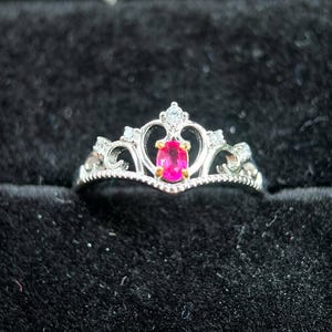 May include: A silver-toned crown ring with a vibrant pink oval gemstone at its center, set within a wooden jewelry box with a black velvet interior. The ring features delicate filigree and small clear accent stones.