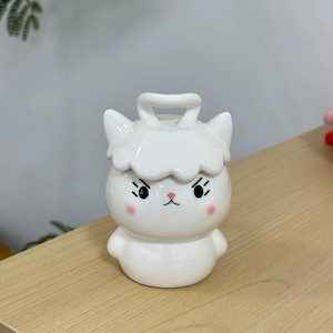 May include: A white ceramic container shaped like a cartoon sheep with a handle on top. The sheep has black eyes, pink cheeks, and a pink nose. The container is on a light brown wooden surface.