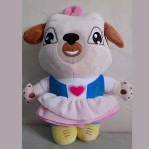 May include: A plush toy dog with a white face, brown ears, and a pink bow. The dog wears a blue vest, a pink skirt, and yellow shoes. A pink heart is on the vest.