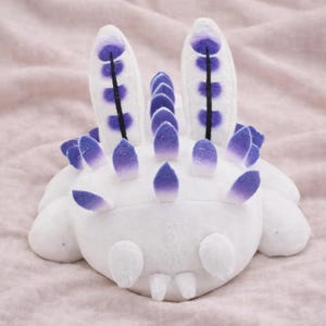 Pode incluir: Um brinquedo de pelúcia de lesma do mar branca com detalhes roxos. O brinquedo tem duas apêndices grandes e verticais com detalhes roxos e uma fileira de espinhos roxos nas costas. A lesma tem um corpo arredondado.