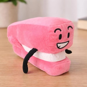 May include: A plush pink stapler toy with a smiling face, black arms, and a white base. The toy has a cartoonish design and is made of soft fabric. The stapler's mouth is open, revealing a white interior.
