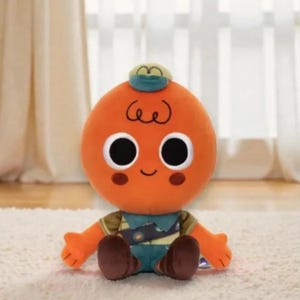 May include: Orange plush toy with large black eyes, a small smile, and brown feet. The toy has small arms and a green and blue outfit. A small green hat sits atop the orange head. The toy is sitting on a white surface.