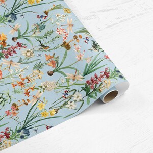 May include: A roll of wallpaper featuring a detailed botanical design. The pattern includes various flowers, dragonflies, and birds in shades of red, orange, yellow, and white against a light blue background. The paper is rolled up, showing the pattern's repeating nature.