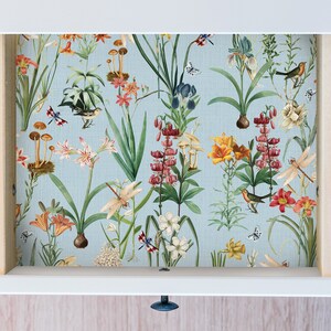 May include: A drawer interior featuring a floral wallpaper design. The wallpaper has a light blue background with illustrations of various flowers, mushrooms, birds, and butterflies. The drawer is white with a wooden frame.
