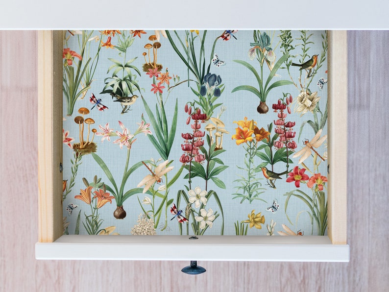 May include: A drawer interior featuring a floral wallpaper design. The wallpaper has a light blue background with illustrations of various flowers, mushrooms, birds, and butterflies. The drawer is white with a wooden frame.