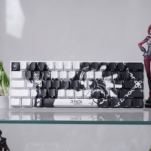 May include: A mechanical keyboard featuring a black and white graphic design across its keys, with Japanese text printed on the spacebar. The keyboard is surrounded by several anime-style character figurines, including figures in dark and red outfits.