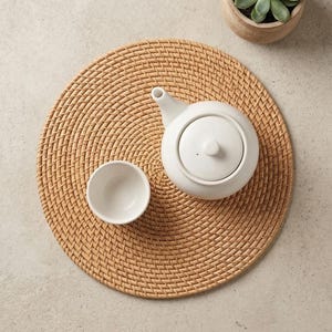 Peut inclure: Une théière et une tasse en céramique blanches sont posées sur un dessous de plat rond en osier brun clair. Une petite plante succulente en pot est dans le coin supérieur droit. La scène est sur une surface texturée neutre.