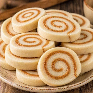 May include: A plate of spiral cookies. The cookies are light beige with a brown swirl in the center. The cookies are stacked on top of each other on a beige plate. The background is a wooden surface.