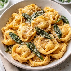 Puede incluir: Primer plano de un cuenco lleno de pasta tortellini. La pasta está cubierta con hojas de salvia fresca, queso rallado y pimienta negra. La pasta es de color amarillo claro y las hojas de salvia son verdes.