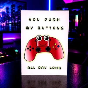 May include: A white card with the text "YOU PUSH MY BUTTONS ALL DAY LONG." The card features a cartoon red game controller with a smiling face. The background is a blurred image of a computer setup.