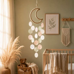 Può includere: Una giostrina sospesa con una forma a mezzaluna avvolta in corda color marrone chiaro, con molteplici dischi circolari traslucidi nelle tonalità del rosa pallido, verde e bianco, appesi alla luna tramite sottili fili bianchi a cascata.