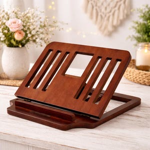 May include: A wooden book stand with a rich, dark brown finish. The stand features a slatted design with a rectangular cutout and a lower lip to hold books. The stand is adjustable and designed to hold books or tablets.
