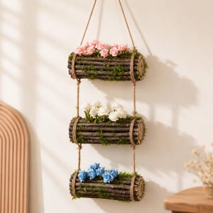 May include: A three-tiered hanging wall planter made of dark brown cylindrical wooden logs covered in green moss. Each tier is suspended by thick jute rope and contains artificial flowers: pink roses on top, white roses in the middle, and blue roses at the bottom.