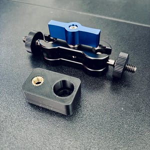 May include: A black metal articulating arm with a blue T-shaped tightening knob and two threaded ends, accompanied by a small black rectangular mounting block featuring a single brass-colored threaded insert and a circular opening.