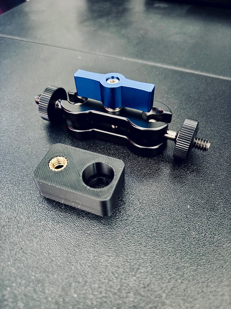 May include: A black metal articulating arm with a blue T-shaped tightening knob and two threaded ends, accompanied by a small black rectangular mounting block featuring a single brass-colored threaded insert and a circular opening.