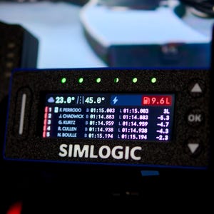 May include: A black Simlogic digital racing dashboard display featuring a rectangular screen with white text showing race standings, lap times, and fuel levels. The device has five green LED indicator lights along the top edge and navigation buttons.