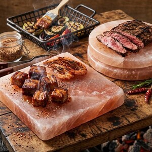 Op de afbeelding: Een culinaire scène met gegrild vlees en groenten. Gesneden steak ligt op een roze zoutblok, naast gegrilde garnalen en varkensvlees. Een klein draadmandje bevat gegrilde groenten en een potje kruiden is zichtbaar.