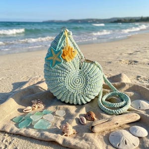 Puede incluir: Bolso de mano de ganchillo azul claro con forma de concha, con una correa larga, decorado con una estrella de mar y un pez. El bolso está en una playa de arena con conchas y vidrio marino. Un accesorio único.
