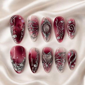 Pode incluir: Um conjunto de dez unhas postiças em formato amendoado com um degradê de bordô profundo e branco translúcido. Cada unha é decorada com relevos metálicos prateados em 3D estilo líquido, incluindo asas de borboleta, um coração e espirais.