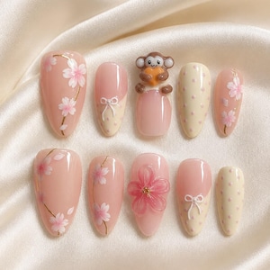 Puede incluir: Un juego de diez uñas postizas con acabado brillante en tonos rosa pálido y crema. Los diseños incluyen flores de cerezo pintadas a mano, lazos blancos en 3D, lunares crema y una figura de mono marrón en relieve que sostiene un corazón.
