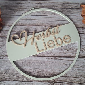 Pode incluir: Uma decoração de parede circular com uma moldura verde sálvia clara e uma barra horizontal central. As palavras "Herbst Liebe" estão escritas num tom de madeira claro, com uma forma de coração integrada na primeira letra do texto.