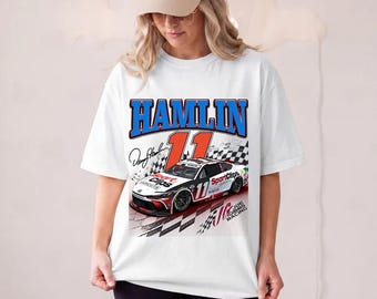 May include: A white short-sleeve t-shirt featuring a graphic print of a race car with the number 11, the name HAMLIN in blue letters, a signature, and the text SportClips and Joe Gibbs Racing. The design includes a checkered flag pattern.