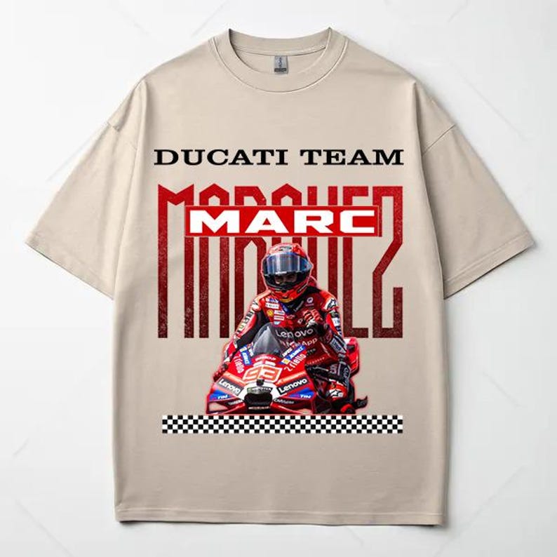 May include: A beige short-sleeve t-shirt featuring a graphic print of a motorcycle racer in a red helmet and suit on a red Ducati motorcycle. The text DUCATI TEAM and MARC MARQUEZ appears in black and red, with a black and white checkered border below.