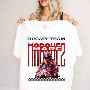 May include: A white t-shirt featuring a graphic of a motorcycle racer in a red and white suit on a red motorcycle. The text reads DUCATI TEAM and MARC MARQUEZ in bold black and red lettering. A black and white checkered pattern is at the bottom.