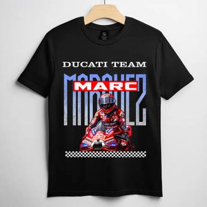 May include: A black short-sleeve t-shirt featuring a graphic print of a motorcycle racer in a red suit on a red Ducati motorcycle. The text reads DUCATI TEAM, MARC, and MARQUEZ in white and red, with a black and white checkered pattern at the bottom.
