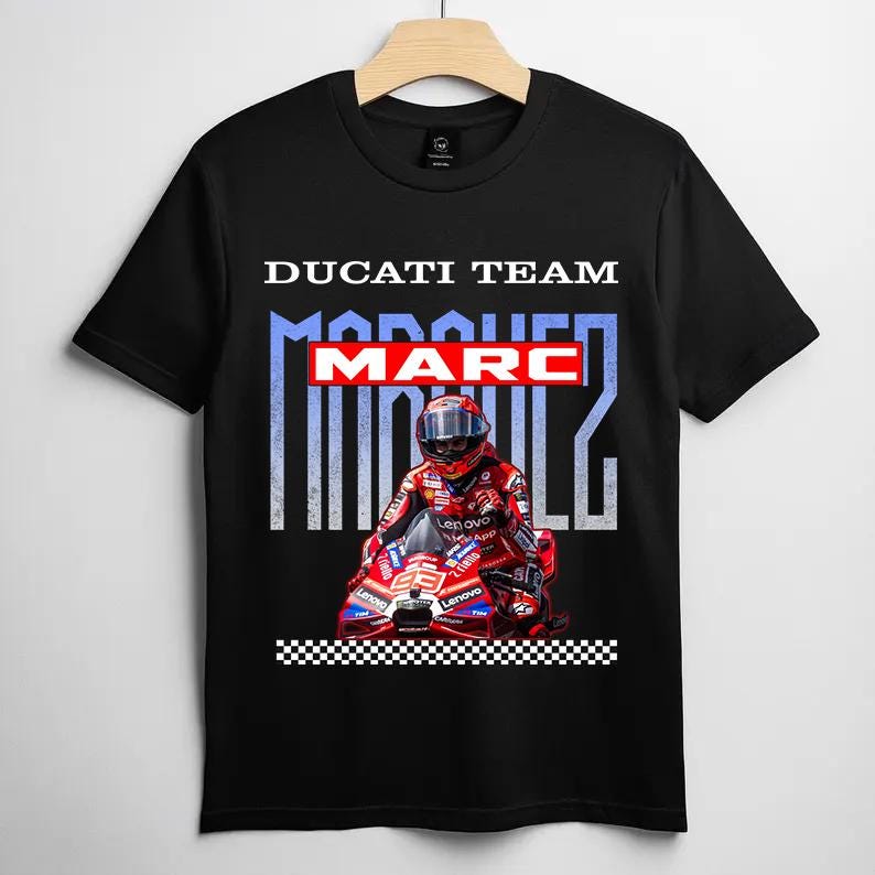 May include: A black short-sleeve t-shirt featuring a graphic print of a motorcycle racer in a red suit on a red Ducati motorcycle. The text reads DUCATI TEAM, MARC, and MARQUEZ in white and red, with a black and white checkered pattern at the bottom.