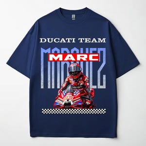 May include: A navy blue short-sleeve t-shirt featuring a graphic print of a motorcycle racer in red gear on a red motorcycle. The text reads DUCATI TEAM, MARC in a red box, and MARQUEZ in large light blue letters, with a black and white checkered border.