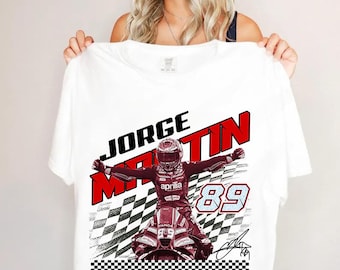 May include: A white t-shirt featuring a graphic print of motorcycle racer Jorge Martin in a racing suit on a motorcycle. The design includes the text JORGE MARTIN in red, the number 89 in blue, a checkered flag pattern, and the Aprilia Racing logo.