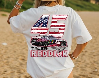 May include: White t-shirt featuring a race car design with the number 45 in red and blue, resembling the American flag. The shirt also displays the name "Tyler Reddick" in red, along with the Toyota logo.
