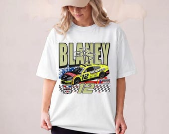 May include: A white short-sleeve t-shirt featuring a graphic print of a yellow number 12 race car with the text Blaney, Menards, and Team Penske. The design includes a checkered flag pattern, blue stars, and a signature across the top section.