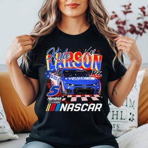 May include: Black t-shirt featuring a graphic of a blue race car with the number 5 and the name "Kyle Larson." The shirt also includes the word "NASCAR" and the Hendrick logo. The design incorporates red, white, and blue colors.