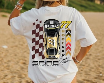 May include: A white t-shirt featuring a graphic on the back with a top-down view of a black and gold race car numbered 77. The design includes a brown checkered pattern, the text "SPIRE MOTORSPORTS", "HOCEVAR", and a signature in black ink.