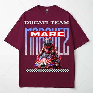 May include: A maroon short-sleeve t-shirt featuring a graphic of a motorcycle racer in a red suit and helmet on a red motorcycle. The text DUCATI TEAM and MARC MARQUEZ appears in white and blue, with a black and white checkered pattern at the bottom.