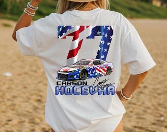 May include: White t-shirt featuring a graphic of a race car with the number 77 and the name "Carson Hocevar". The design incorporates the American flag's stars and stripes. The shirt is a casual wear item.