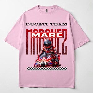 May include: A light pink short-sleeve t-shirt featuring a graphic print of a motorcycle racer in red gear on a red motorcycle. The text DUCATI TEAM and MARC MARQUEZ is printed above the racer, with a black and white checkered pattern at the bottom.