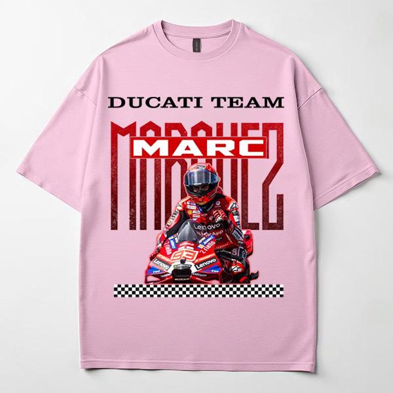 May include: A light pink short-sleeve t-shirt featuring a graphic print of a motorcycle racer in red gear on a red motorcycle. The text DUCATI TEAM and MARC MARQUEZ is printed above the racer, with a black and white checkered pattern at the bottom.