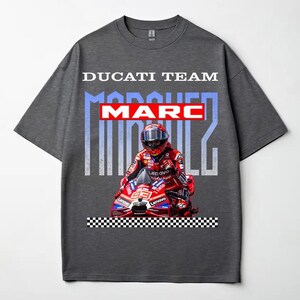 May include: A dark gray short-sleeve t-shirt featuring a graphic print of Marc Marquez on a red Ducati motorcycle. The text DUCATI TEAM appears at the top, with MARC MARQUEZ in large letters behind the rider. A black and white checkered pattern is at the base.