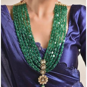 Puede incluir: Un collar de múltiples hilos con cinco filas de cuentas ovaladas de color verde pulido. El collar incluye un colgante central vertical compuesto por tres engastes rectangulares y una base en forma de flor, con piedras claras y metal dorado.