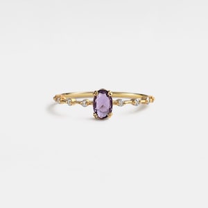 Könnte beinhalten: Ein goldener Ring mit einem ovalen, lila Edelstein in der Mitte. Das Band ist mit kleinen, klaren Edelsteinen verziert. Der Ring ist vor einem weißen Hintergrund platziert, was sein elegantes Design hervorhebt.