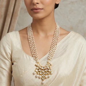 Peut inclure: Un collier de perles à plusieurs rangs avec un pendentif doré. Le pendentif présente des accents clairs et dorés, avec des perles pendantes. Le collier est porté sur un vêtement de couleur crème.