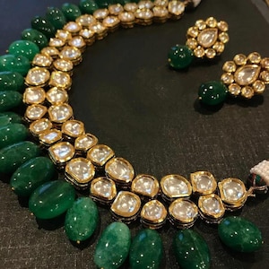 Könnte beinhalten: Ein goldfarbenes Halsketten- und Ohrring-Set. Die Halskette hat Reihen aus smaragdgrünen Perlen und goldgerahmten, klaren, facettierten Steinen. Die passenden Ohrringe haben ein goldenes Blumendesign mit einem grünen Perlenanhänger.