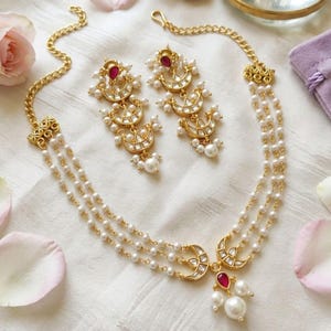 May include: A gold-toned necklace and matching earring set featuring three strands of small white pearls. The jewelry includes crescent-shaped metal accents, clear stones, and teardrop-shaped red stones, with a dangling pearl at the center.